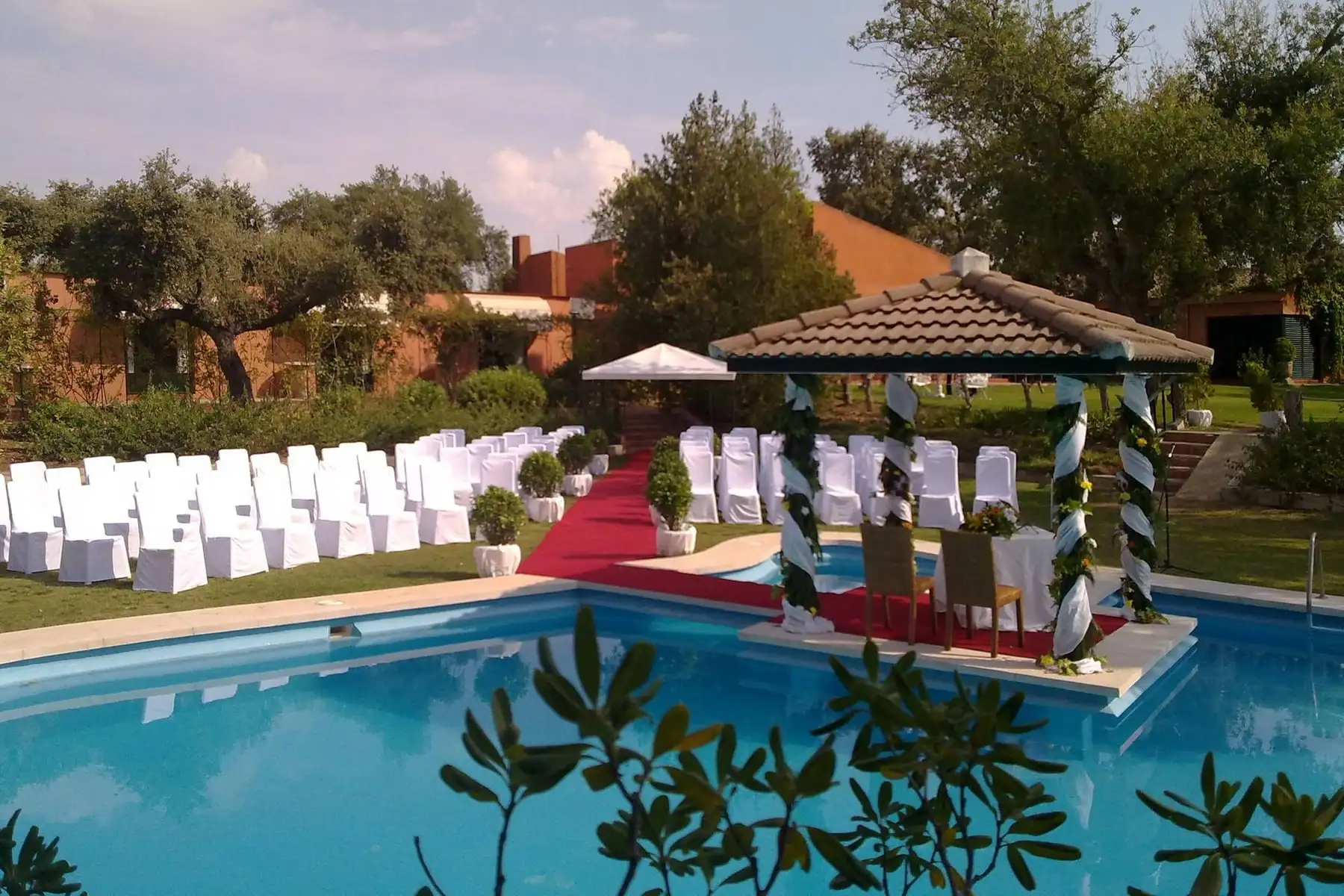 La ceremonia civil preparada: sillas blancas sobre el césped, una alfombra roja y un templete con flores sobre la piscina