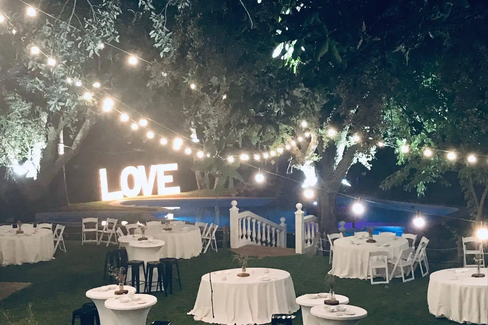 Mesas redondas y altas sobre el césped bajo guirnaldas de luces, con las letras LOVE y la piscina al fondo
