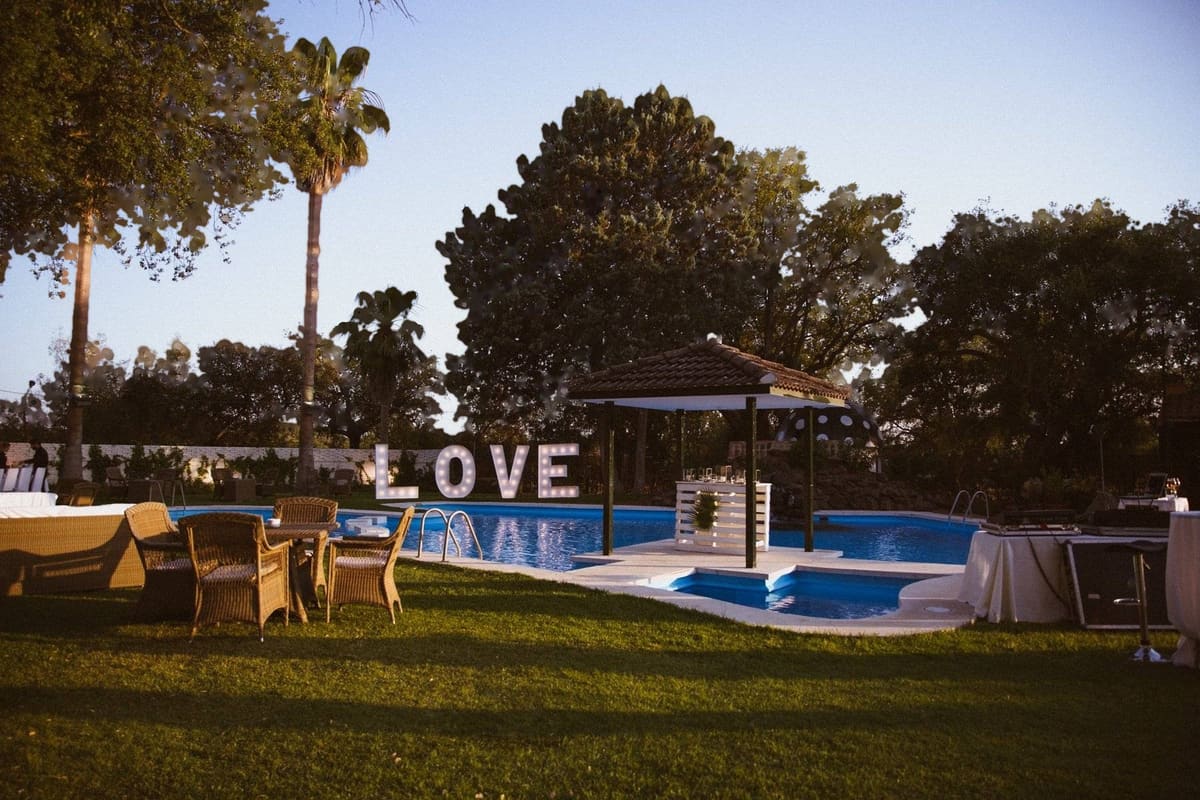 El jardín del cortijo al atardecer: la piscina con su templete y las letras LOVE, un gran árbol y palmeras bajo un cielo azul